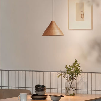 "The Luma wooden pendant light, with its modern pyramid shape, hangs above a dining table set with bowls, plates, and a glass vase with greenery, casting a soft, ambient light in the contemporary space."