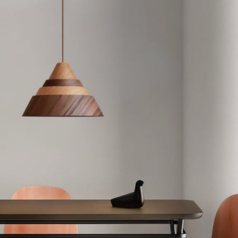 "The Luma wooden pendant light, with its distinctive pyramid-like structure and varying wood tones, hangs above a table with a black bird ornament, providing a soft, ambient glow in a minimalist setting."