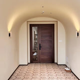 "Two travertine wall lamps with cylindrical shades and wooden bases, illuminating a hallway with a patterned floor and a dark wood door."