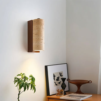 "A modern wall lamp with a cylindrical travertine shade and a wooden base, casting a warm glow in a minimalist room."