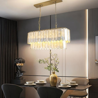 "Linear chandelier with scalloped shell panels and gold accents, hanging above a dining table in a modern dining room with a bust sculpture."