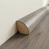 Rounded wooden trim with a light gray finish, displaying a smooth surface and angled back edge against a floor and wall.