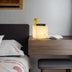 Abak alabaster table lamp with a cube shape, warm glow, yellow vase with flowers, and open book on a wood bedside table.