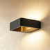 Black square wall sconce with gold interior detail mounted on white concrete exterior wall with blurred architectural backgro