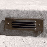 "Close-up of a brass step light fixture with horizontal grille design, made from durable, weather-resistant brass, installed on a concrete step for outdoor lighting"