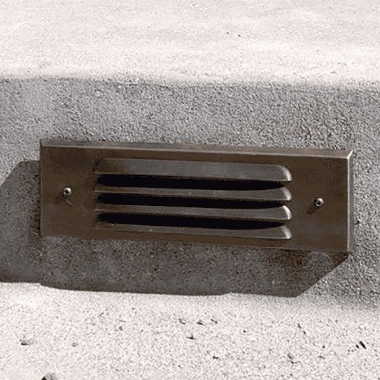 "Close-up of a brass step light fixture with horizontal grille design, made from durable, weather-resistant brass, installed on a concrete step for outdoor lighting"