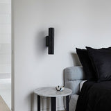 "Black travertine cylindrical wall lamp mounted on a bedroom wall beside a bed and side table."