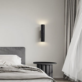 "Black travertine cylindrical wall lamp mounted on a bedroom wall beside a bed and curtains, casting light upwards and downwards."