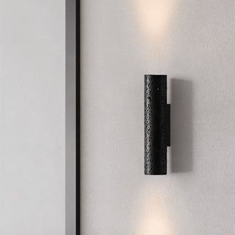 "Black travertine cylindrical wall lamp mounted on a light wall, casting light upwards and downwards."