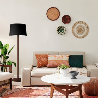 "A modern floor lamp with a black drum shade and a slender wood stand complements a boho-style living room with a neutral sofa, patterned pillows, woven wall decor, and a round coffee table."