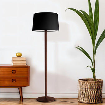 "A modern floor lamp with a black drum shade and a slender wood stand stands next to a wooden dresser and a potted plant in a room with a white wall."