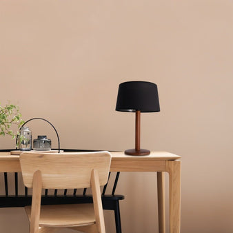 "A contemporary table lamp with a black fabric shade and a wood-textured metal base sits on a light wood desk in a minimalist office setting with a light beige wall."