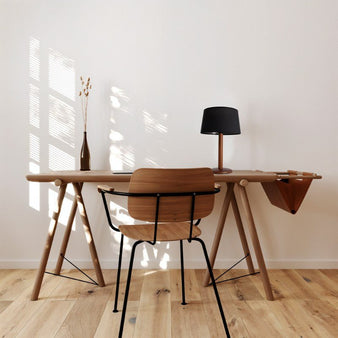 "A contemporary table lamp with a black fabric shade and a wood-textured metal base, placed on a light wood desk in a modern office with a wooden chair."