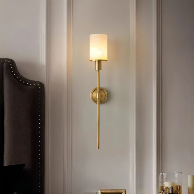 "A brass wall sconce with a cylindrical alabaster shade mounted on a light gray wall next to a bed."