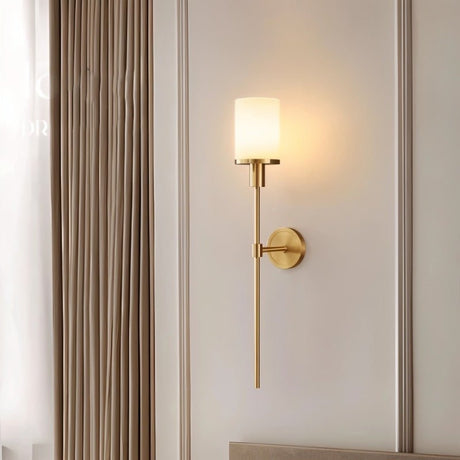 "A brass wall sconce with a cylindrical alabaster shade casting a warm light in a modern bedroom, with a curtain visible on the left."