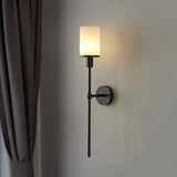 "A modern wall lamp with a cylindrical alabaster shade, mounted on a wall next to a dark curtain, providing soft lighting in a bedroom."