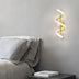 A stylish wall lamp with a gold finish and a twisting light design, installed beside a cozy bed with gray bedding.
