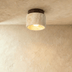 Cylindrical travertine pendant light with warm brass top mounted on white ceiling in hallway with wood paneling and green pla