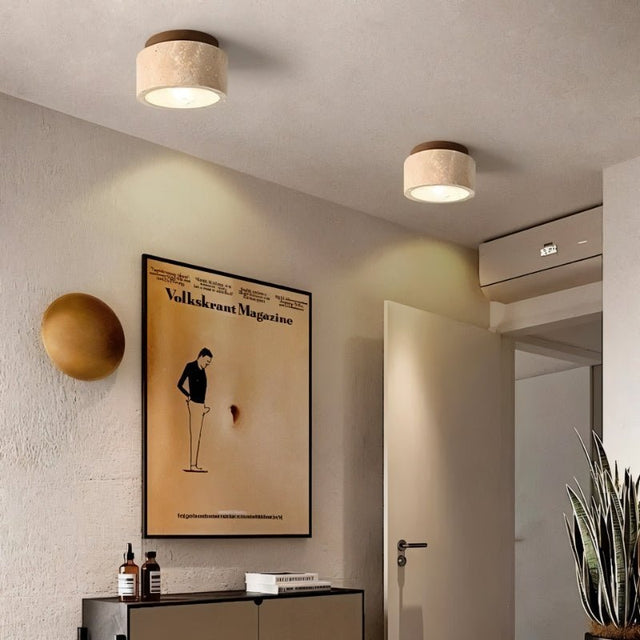 "Two cylindrical yellow travertine ceiling lights with dark wood tops in a hallway with artwork, modern interior lighting."