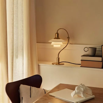 "Alya Table Lamp with a curved metallic arm and frosted glass shades, made from aluminum, iron, and glass, placed on a desk with books and a cup, offering soft, diffused lighting."