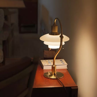 "Alya Table Lamp featuring a curved metallic arm and frosted glass shades, made of aluminum, iron, and glass, placed on a side table with books, casting soft, diffused lighting."