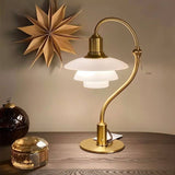 "Alya Table Lamp with a curved metallic arm, frosted glass shades, and a brass finish, placed on a wooden table with decorative objects, providing soft, diffused lighting."