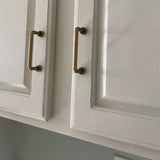 "Two minimalist antique brass pull bar handles made of durable zinc, installed on white cabinets, showcasing a slim, cylindrical design with rounded ends."