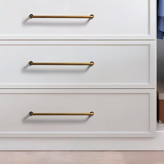 "Three minimalist antique brass pull bar handles made of durable zinc, installed on white dresser drawers, showcasing a slim, cylindrical design with rounded ends."