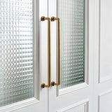 "Two long, minimalist antique brass pull bar handles made of durable zinc, installed on white framed glass doors, featuring a slim, cylindrical design with rounded ends."