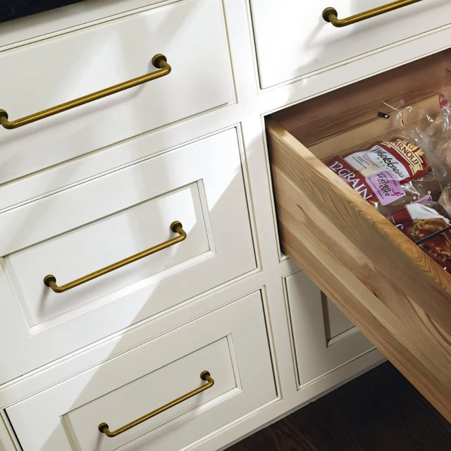 "Minimalist antique brass pull bar handles made of durable zinc, installed on white kitchen drawers, one of which is open and filled with bread."