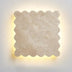 "Close-up of the square Amon travertine wall lamp, showcasing its distinctive scalloped edges and the natural variations in the travertine stone, illuminated from behind with a soft, warm light."