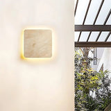 "A square Amon travertine wall lamp with a smooth, minimalist design is mounted on a light-colored wall, illuminating a modern outdoor patio with a metal pergola, lush green plants, and a glimpse of a white building."