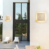 "Two square Amon travertine wall lamps with a smooth, minimalist design illuminate the exterior of a modern house with large windows, a black framed door, and a spacious patio with white lounge chairs."