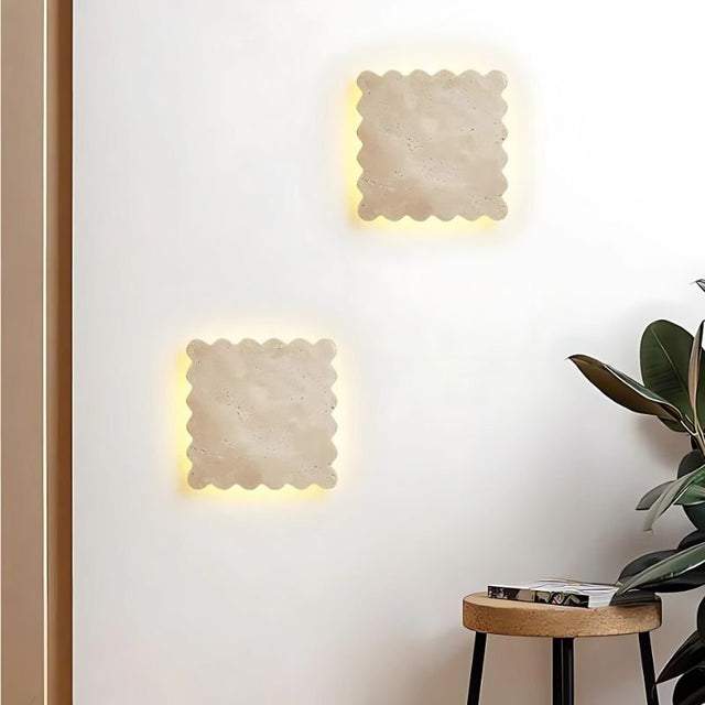 "A pair of square Amon travertine wall lamps, each with scalloped edges, are symmetrically placed on a light-colored wall, creating a modern and elegant ambiance above a small round table with a book."