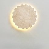 "Close-up of the Amon travertine wall lamp, showcasing its unique scalloped, flower-like design and the natural texture of the travertine stone, illuminated by a soft, warm glow."