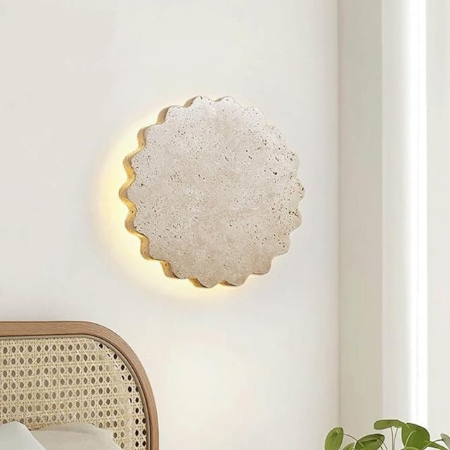 "The Amon travertine wall lamp, with its scalloped, flower-like design, installed on a light-colored wall, casting a warm glow in a room with a rattan chair."