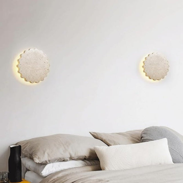 "A pair of Amon travertine wall lamps, with a scalloped, flower-like design, flank a minimalist bed with layered pillows and a dark vase, creating a serene and modern bedroom ambiance."
