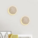"A pair of Amon travertine wall lamps, with a scalloped, flower-like design, symmetrically installed on a light wall, creating a warm and elegant ambiance above a bed with a yellow textured throw."