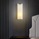 "A modern, rectangular alabaster wall lamp with a subtle ambient glow, framed by a thin, dark metal rod and brass accents, mounted on a dark textured wall in a bedroom setting."