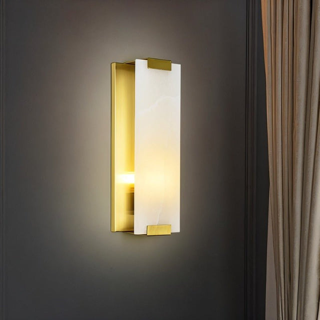 "A modern wall sconce featuring a rectangular alabaster panel illuminated from within, framed by brass accents, mounted on a dark gray wall."