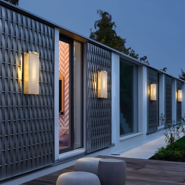 "Several modern wall lamps with rectangular alabaster panels and metal frames, illuminating the exterior of a building with a textured facade at dusk."
