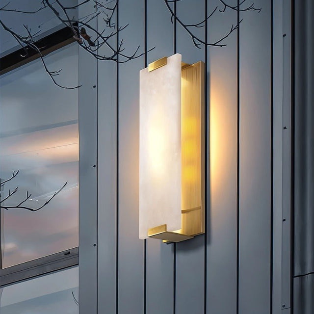 "A modern outdoor wall sconce with a rectangular alabaster panel and brass frame, casting a warm glow on a textured exterior wall at dusk."