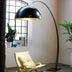Tall arc floor lamp with a black dome shade and gold interior, illuminating a cozy seating area with plants and decor.