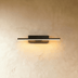 Black metal angular wall sconce with illuminated linear rod, mounted on white wall above wooden bed frame and pink flowers.
