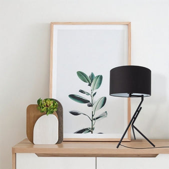 "Anubi table lamp with a black fabric shade and geometric base on a dresser in a modern bedroom setting with a plant picture frame."