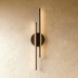 Black modern wall sconce with horizontal bars and circular center element, mounted above bedroom nightstand in warm lighting.