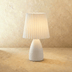 Pink ceramic table lamp with pleated shade on white surface beside pink flowers and gray sofa, studio setting.