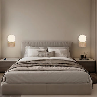 "A pair of modern wall lamps with spherical glass shades and travertine bases, flanking a contemporary bed with soft, ambient light."