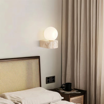 "A modern bedroom featuring a spherical wall lamp emitting a soft glow, mounted on a textured travertine base beside a rattan headboard."