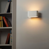 "White gypsum Arcat wall lamp with a rounded rectangular shape, emitting soft light on a wall near a bookshelf."